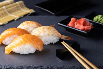 Sushi Sake with salmon and Ebi with tiger shrimp in focus on a black slate board on a dark background, with ginger, wasabi, soy sauce in a blur and chopsticks. The concept of a menu of Asian restauran
