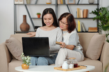 Smiling senior Vietnamese woman paying for online purchases of her adulr daughter when they are drinking tea in living room together