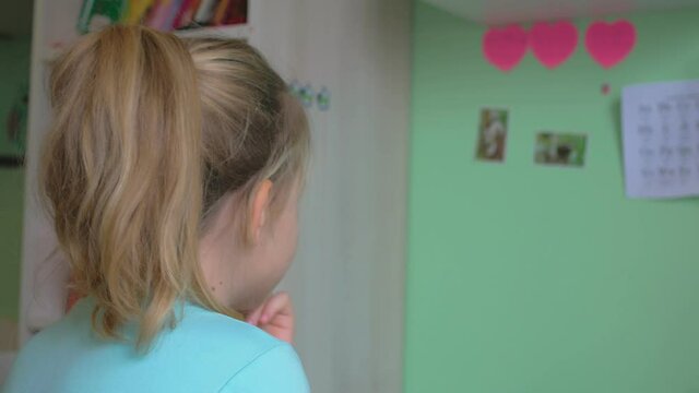 In a room with gray-green wallpaper, a girl with fair hair sits, thrusting her fingers into her mouth with excitement, there are three red paper hearts on the wall. back view