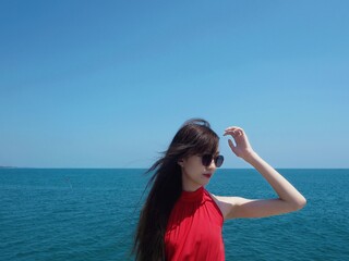 Asian lady in red dress, she ware sunglass sit on the bridge while she raised a hand against the blown hair, the sea breeze blew her hair