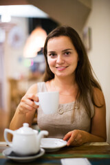 Portrait of cheerful young girl rests in cafe behind cup of tea