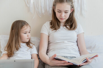 A little girl with a laptop looks into her older sister's book. The concept is a fascination with reading or tablet.