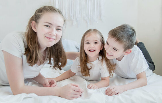 The concept is happy brother and sister. Three children of different ages: a teenage girl, a little girl and a boy. Children in white T-shirts lie on the white bed and smile.