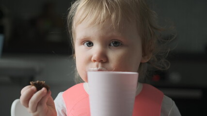 Front view of little cute white girl blonde with pink bib drinks kefir from pale pink glass and...