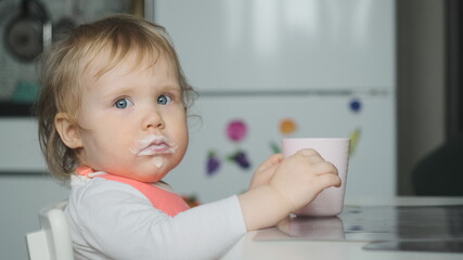 Side view little cute white girl blonde with pink bib drinks kefir from pale pink glass and looks at camera. Mouth in kefir or yogurt. Baby wear white shirt or bodysuit.