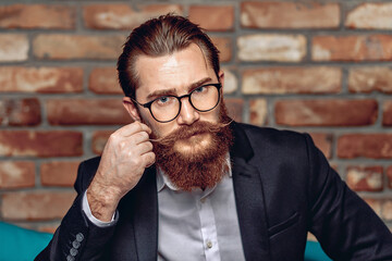 Portrait of a charismatic brutal young man with a mustache and beard holding glasses and posing against a background of a brick wall. Male charisma concept