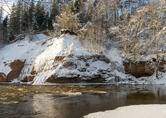sunny day, landscape with red sandstone cliffs that are snowy with snow, frozen icicles on the cliff wall, icefall on the cliff wall, frozen river, Gauja, Kuku cliffs, Latvia