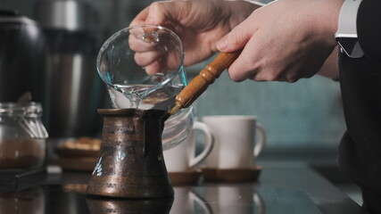 woman prepares coffee in turk on the burner in the kitchen. Lady pours water into a coffee maker. There are white caps in the background