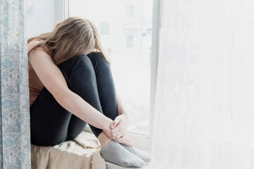 The teenage girl sits on the window sill bending her legs and burying her face in her knees. Concept of depression, loneliness in teenagers.