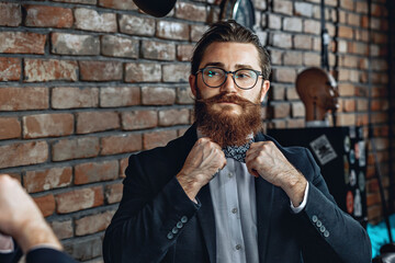 Portrait of an attractive fashionable man with glasses, beard and mustache, looking at himself in the mirror and straightening a bow tie on his collar. Luxury concept