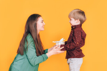 Little boy gives mom a box with a gift against a yellow background. Gift for womens day and mothers day.