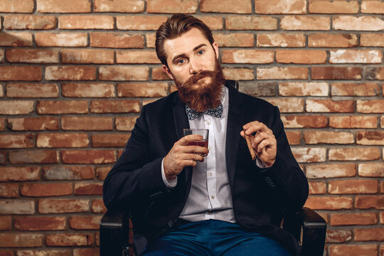 Portrait of a brutal gentle man with a mustache and a beard sitting on a chair and holding a glass of whiskey and a cigar in his hand, posing against a brick wall. Male beauty concept