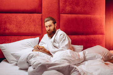 Young well-groomed man with a long mustache and beard, wearing a robe, sitting in bed and holding a cream in his hands. Personal care. Spa concept