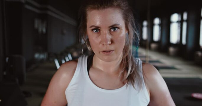 Portrait of serious beautiful young Caucasian female athlete looking at camera, sweaty after intense workout at gym.