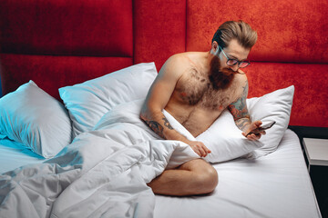 Sexual focused adult man wearing glasses and sitting on a white bed looking at the phone before going to bed. Night time concept