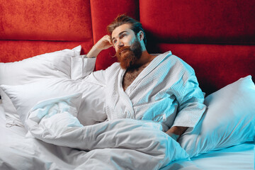 Adult sexual dreaming man wearing white robe and lying in bed under a white blanket looking upward thinking. Male beauty concept