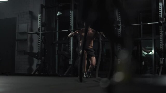 Athletic man performs exercises with battle ropes, endurance training in the gym, cinematic light, 4k slow motion.