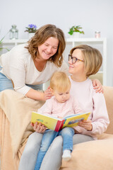 Two young women are sitting in the living room on the couch with a child on their knees and are reading a book to the child. Gay lesbian couple at home with a baby. Lesbian family concept