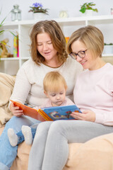 Obraz premium Homosexual family of girls at home with their child. Spouses are sitting on the couch reading a book and looking at their daughter