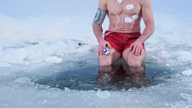 Medical test in the cold. Man shivers with cold while sitting on the ice with legs immersed into icy water. Oximeter, blood pressure monitor and cardio electrodes are connected to his body