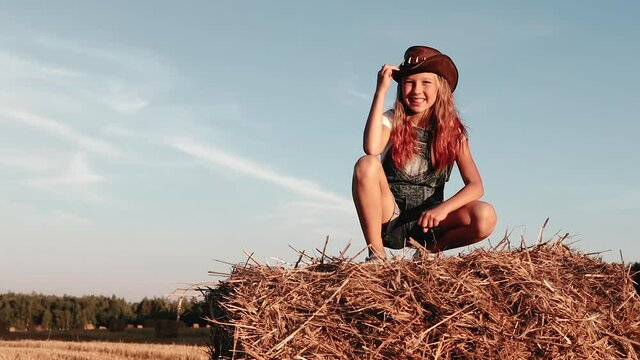 Pretty girl in cowboy hat poses for photo shoot in rural field sitting on bale of hay. Teenager in cowgirl look against blue sky in harvest field