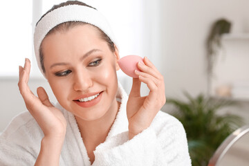 Beautiful young woman with sponge applying makeup at home