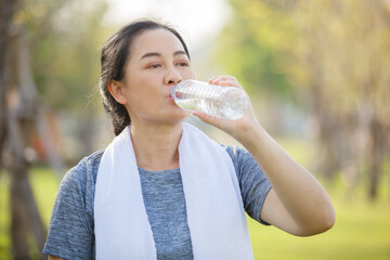 Asian elderly or senior woman drinking water in bottle after exercise for refreshing in garden,.