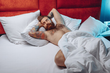 Obraz na płótnie Canvas Top view portrait of sleeping handsome lovely guy with beard mustache and tattoo lying on his back on a white bed and sleeping. Hotel room concept