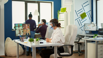 Obraz premium Entrepreneur wearing face mask against coronavirus discussing on video meeting using headset with business partner in new normal financial office. Multiethnic coworkers respecting social distance.