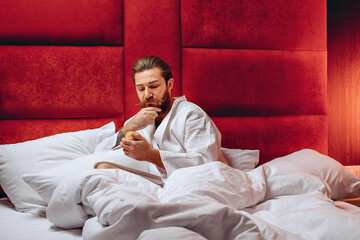 Handsom sharming man with a long mustache and beard, wearing a robe, sitting in bed and holding a cream in his hands. Personal care. Care concept