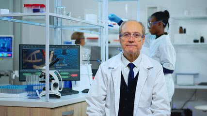 Senior chemist sitting in modern equipped lab looking satisfied at camera. Multiethnic team examining virus evolution using high tech and chemistry tools for scientific research, vaccine development