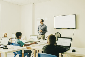 Confident teacher explaining lesson to pupils. Multiethnic children sitting at table in classroom, listening middle-aged man and using laptop computers. Childhood and digital education concept