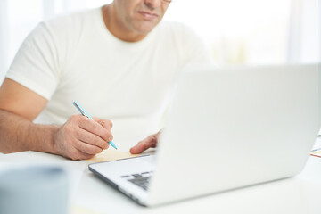 Cropped shot of latin man in white t shirt sitting at the table, using laptop and making notes. Businessman working from home during lockdown