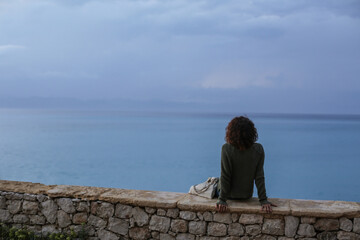 Lifestyle photo of young beautiful woman curly faceless looking at the calm blue sunset Mediterranean sea and dusk cloudy dramatic sky in Rhodes Island, Greece in summer. Soft focus, film edit. 