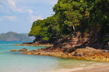 sandy beach with rocks in the foreground, sea turquoise water and tropical vegetation in the background