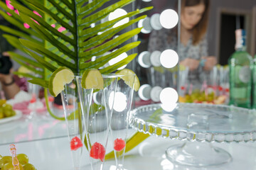 Soft focused close up shot of coctail glasses with berries and lime slices. Plate for cake, lights bokeh, mirror, unrecognizable person on blurry background. Candy bar for festive party.