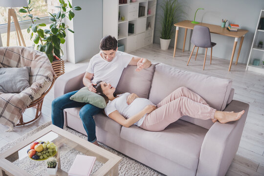 Full size photo of positive young parents sit lie couch talk expect baby enjoy rest indoors in house living room