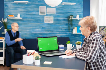 Senior woman taking notes on notebook looking at portable computer with copy space available. Elderly woman working on laptop with green screen and husband holding tv remote control.