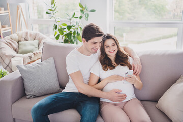 Obraz premium Portrait of tender cheerful couple waiting baby sitting on divan using device browsing blog app in light house flat indoor