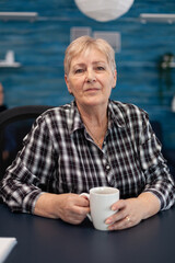 Portrait of senior woman looking at camera while enjoying a mug of coffee. Healthy happy elderly woman in home living room enjoying her retirement