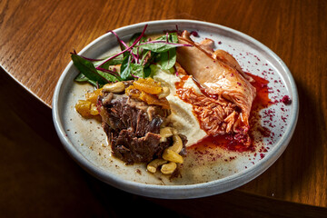 Stewed beef steak with dried fruits and nuts garnished with celery puree and kimchi cabbage, served in a white plate on a wooden background. Restaurant food