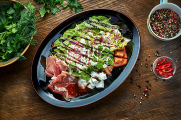 Delicious and dietary salad with jamon, feta cheese, tomatoes, served in a black plate on a wooden background. Restaurant food