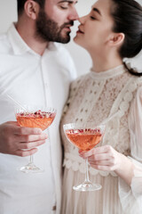 Happy romantic young caucasian couple celebrating, hugging, holding glasses of sparkling champagne cocktail with rose petals and glass drinking straws. Wedding anniversary, Valentines Day concept