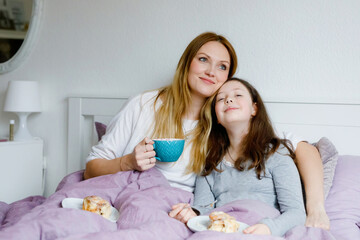 Lovely young mother and cute school kid girl cuddling together in bed in morning. Happy family of young single woman and beautiful daughter having breakfast, eating on mother's day.