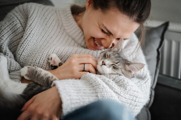 Beautiful cheerful young woman with a cute gray cat in her arms at home on the sofa, friendship and...