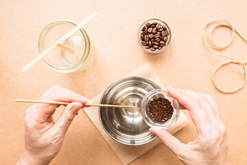 DIY concept. A woman's hand adds ground coffee to wax. Wax, wood wick, coffee are ingredients for making handmade candles. Flat lay. View from above.