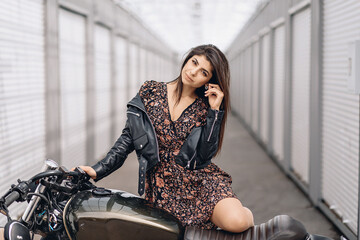 Fototapeta na wymiar Portrait of an adorable sexual model wearing a black leather jacket and posing next to a motorcycle playfully touching her hair. Sensuality concept