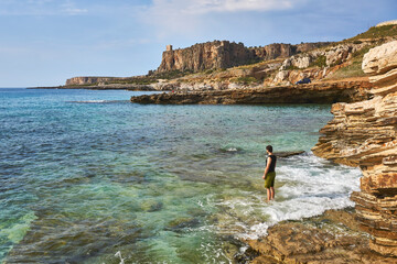 young caucasian man standing with feet int the mediterrenian sea near San vito lo Capo in Sicily looking toward the distance with beautiful coastline in the background