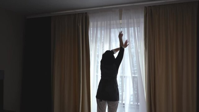 The housekeeper opens the curtains at the hotel room.