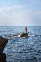 young caucasian man standing on a rock looking at the vast mediterrenian sea on sunny day